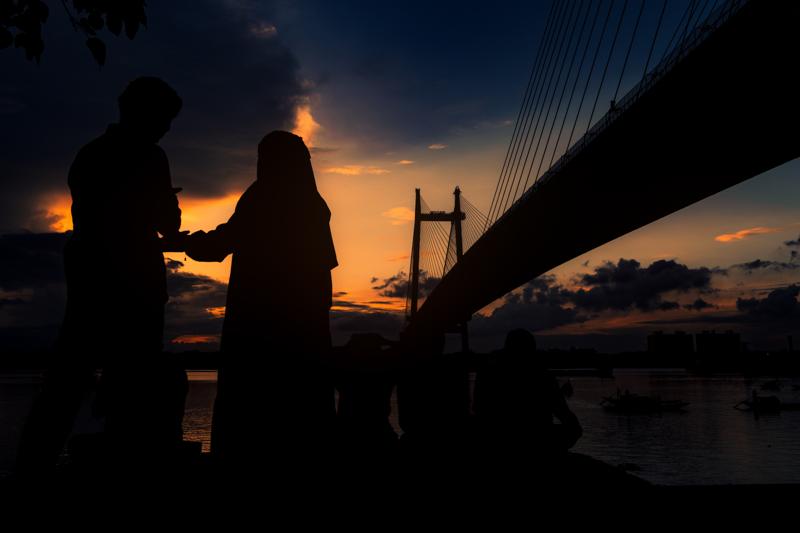 A person stands in silhouette near the still water as the orange horizon and steel bridge lights draw a quiet line behind them.