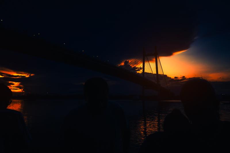 Dark figures stand beside the river as the bridge crosses an orange sunset, its cables and shadowed structure reflected across the broad water.
