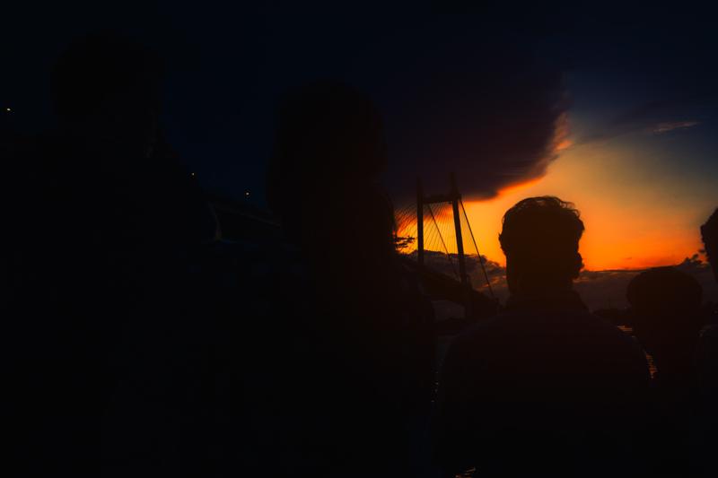 A dark figure stands in profile against the orange sunset, while the riverbank and bridge structure disappear into shadow around them at evening.