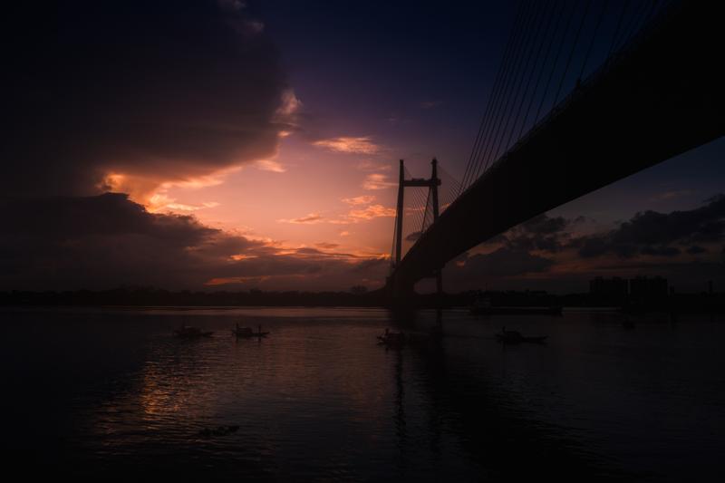 The steel bridge’s cables cut into the blue dusk above scattered riverside figures, with a strip of orange light mirrored on the water.