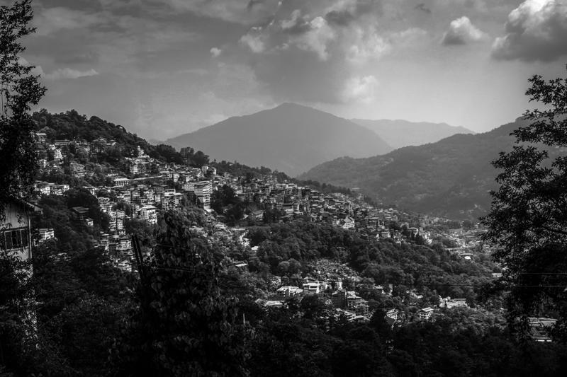 The monochrome view layers a dense hillside settlement beneath distant ridges, while a heavy cloud breaks open above the valley.