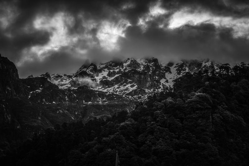 A mountain ridge disappears beneath a sweep of cloud, with the black and white tones sharpening snow, rock and shadow.