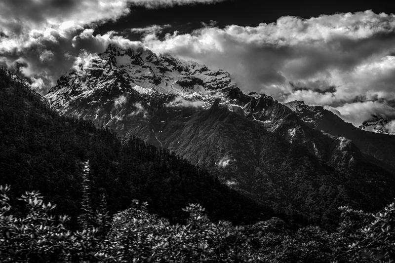 The monochrome valley opens toward distant peaks, their outlines repeatedly hidden and revealed by thick bands of low cloud.