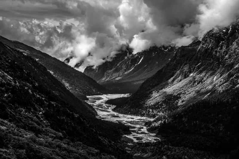 The pale river winds through a broad mountain valley, with steep slopes and scattered cloud enclosing the distant snowline.
