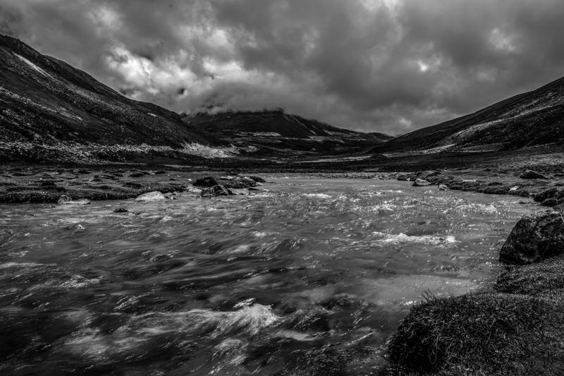 White water cuts through dark rocks in the monochrome frame, leading from the close foreground toward the open valley.
