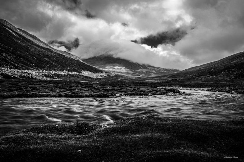 The monochrome river breaks over rocks beneath a wide mountain basin, with steep ridges closing the scene on either side.
