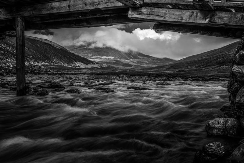 White water runs through a broad rocky valley, framed by dark slopes beneath cloud across the mountain landscape in monochrome.