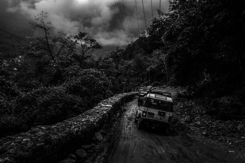 A bus follows the narrow hillside road into drifting mist, with wet rock and dense forest closing around its route.