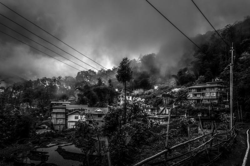 Scattered houses cling to a hillside beneath thick cloud; one tree rises above the roofs while the valley disappears below.