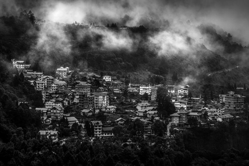 The monochrome hillside town appears in layers, rooftops and a tree emerging from cloud that drifts across the valley below.