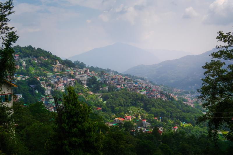 Colourful homes cling to the steep hillside beneath soft cloud, while the green valley fades toward a blue-grey mountain line.