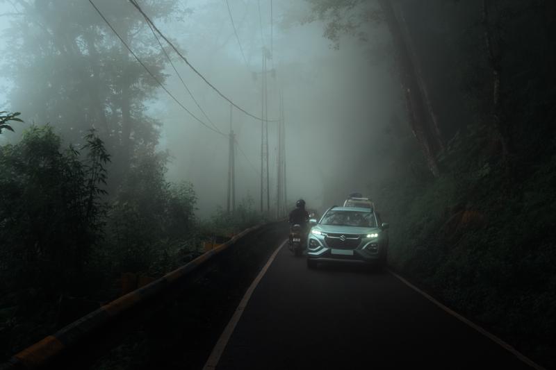 A car rounds misty road toward the camera, its lamps cutting through wet air between the trees and power lines.