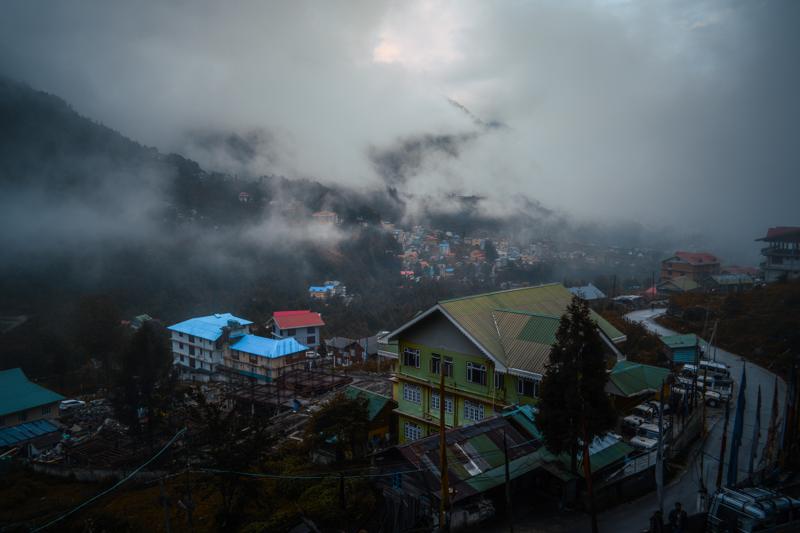 A handful of rooftops appears among the steep green slopes, partly hidden by drifting cloud and linked by the road.
