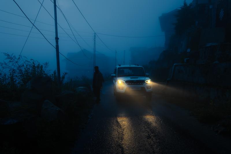A car’s lamps emerge from the fog on a road, with wet trees and wires fading quickly into the haze.