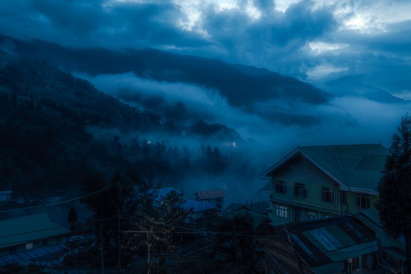 A hillside settlement rests beneath a band of mist, the muted roofs and green slope softening toward the distant ridge.
