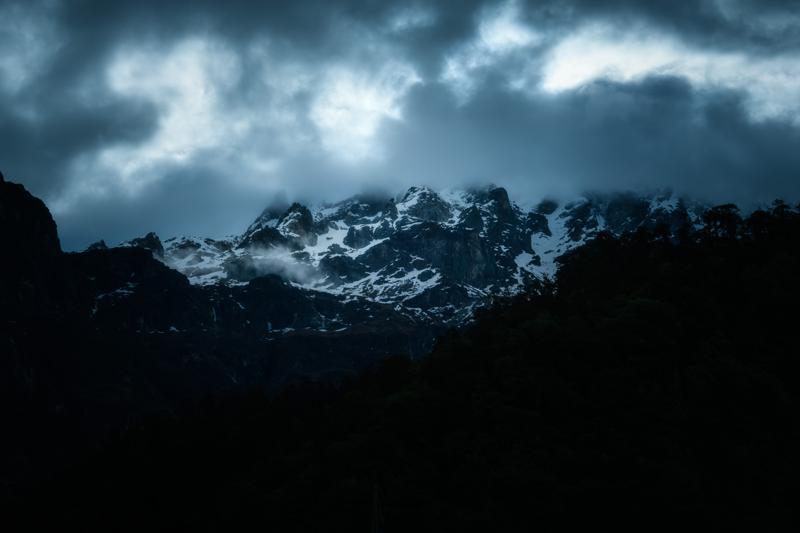 Bright cloud parts over the black mountain ridge, exposing a line of snow and rock above the deep forested foreground.