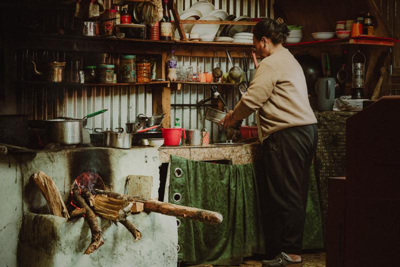 A man stands at a counter lined with jars and goods, while another seated figure and hanging items fill the shop interior.