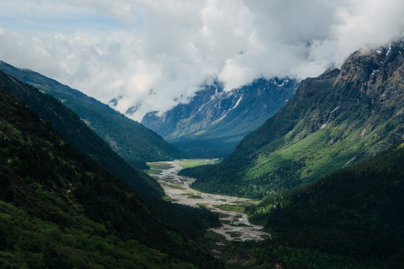 A shallow stream crosses the mountain floor, with stony banks, low grass and distant ridges opening beneath the clouded sky.