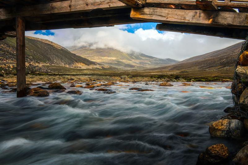 A mountain stream rushes beneath a low wooden beam, with grassy banks and a wide valley visible beyond the water.