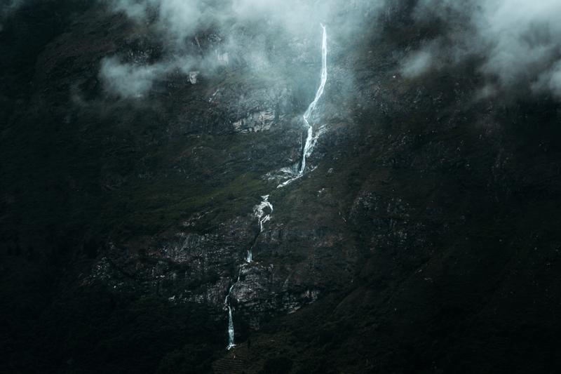 A narrow white cascade drops down the dark rock face, disappearing into dense foliage as mist softens the wooded valley.