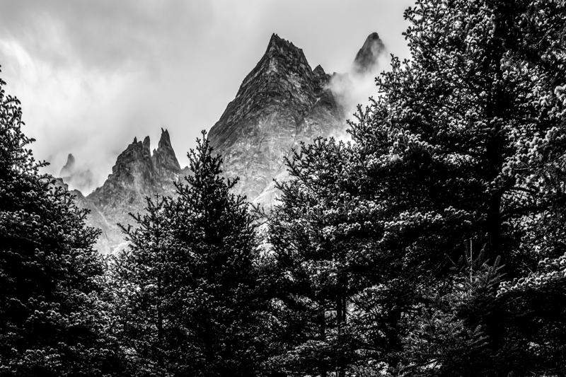 Sharp mountain peaks rise above a dense black forest, while cloud breaks across the ridgeline and opens the monochrome sky.