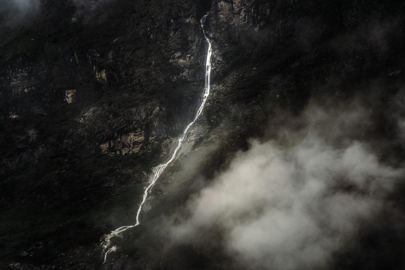 The waterfall descends as a pale thread through the black ravine, its course briefly visible between mist and dense trees.