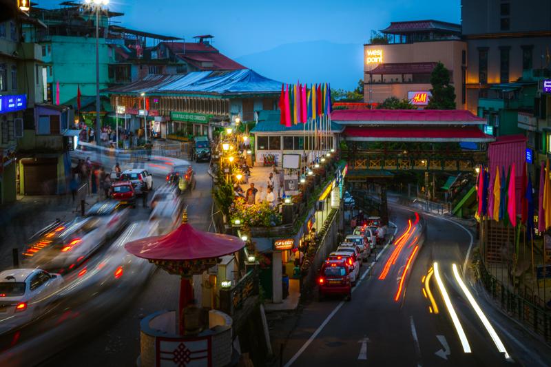 Headlights streak through the hill street, where shop signs and wet blue pavement lead uphill beneath the snowy mountain skyline.
