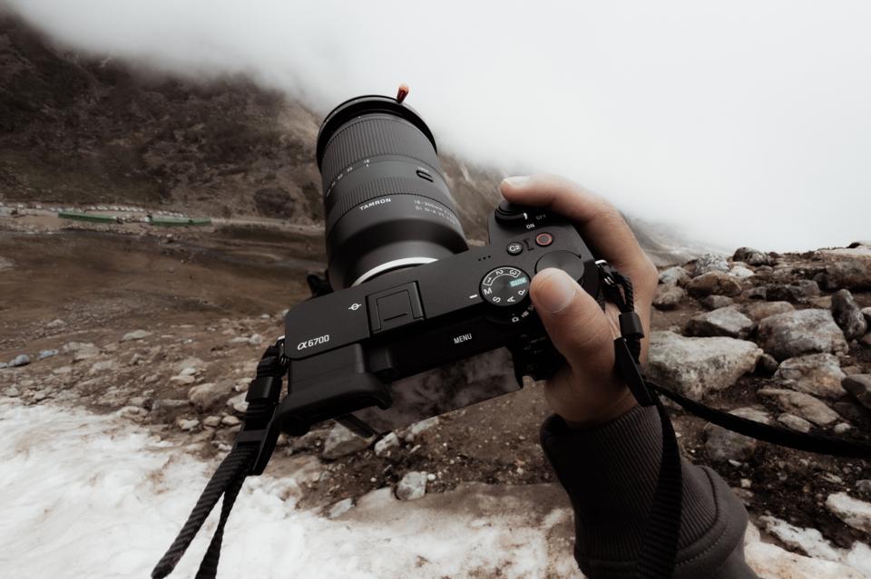 A photographer holds a camera with a telephoto lens above a snowy, rocky mountain landscape.