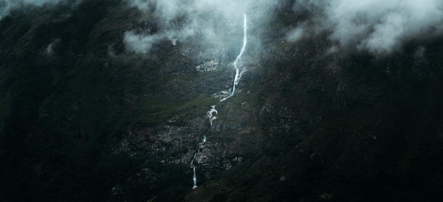 A narrow waterfall flows down a dark mountain beneath drifting clouds and mist.