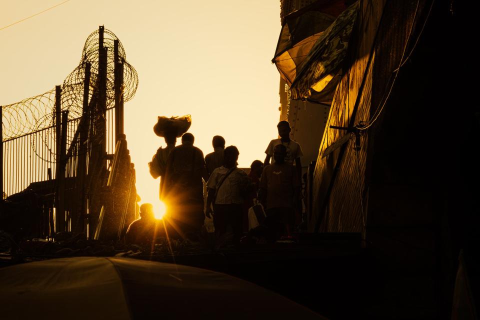 Pedestrians move in silhouette through a narrow passage toward the setting sun.